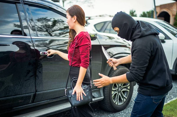 Бандит держит нож, пытаясь ограбить деньги и причинить боль женщине, когда она открывает дверь автомобиля на парковке . Стоковое Фото