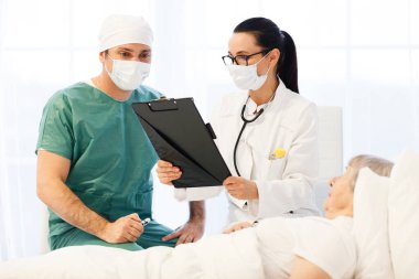 Hastanede sağlık hizmeti. Erkek cerrah ve kadın doktor ya da hemşire yaşlı bayanın yatakta yatıp tedavi olduğunu muayene ediyorlar.