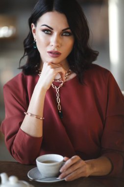 Modern mücevherler içinde güzel esmer iş kadını ve cafe 'de öğle yemeği yiyen şık bir bordo elbise. Kadın masaya oturur ve elinde bir fincan çay tutar.