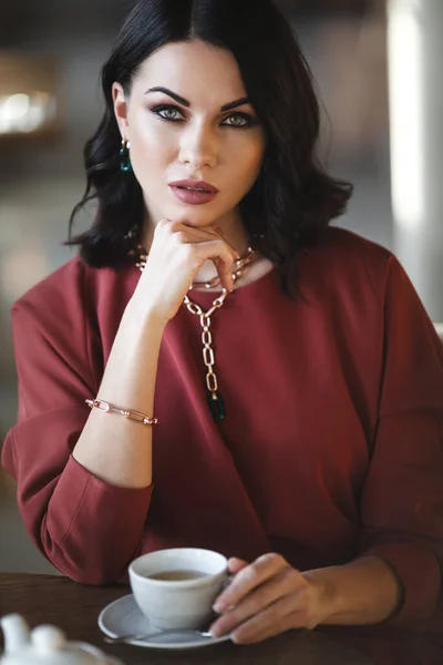 Mooie brunette zakenvrouw in moderne edelstenen en modieuze bordeaux jurk met een lunch in cafe. Vrouw zit aan tafel en houdt een kopje thee vast — Stockfoto