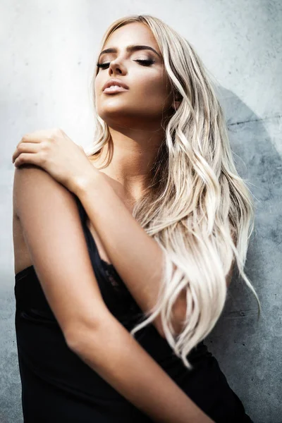 Portret młodej blondynki z pięknym makijażem i włosami w klasycznej czarnej sukience stojącej przy szarej ścianie. Modelka pozowanie w eleganckich ubraniach na świeżym powietrzu — Zdjęcie stockowe