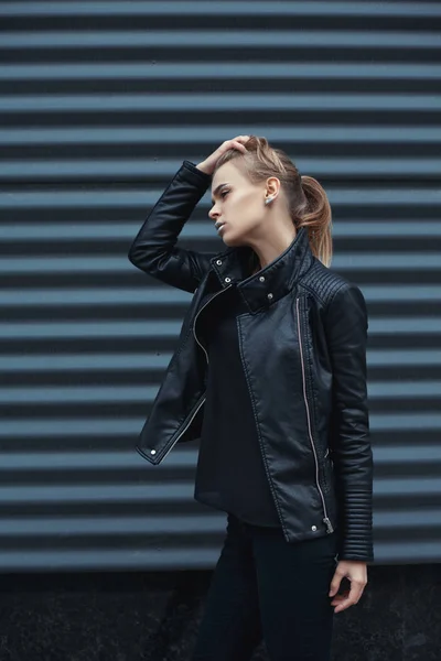 Fashion portrait of young elegant fashionable blonde girl wearing black rock style posing outdoors against urban style background of metal strips. Cold season. Stylish Makeup. Autumn Vogue Style.