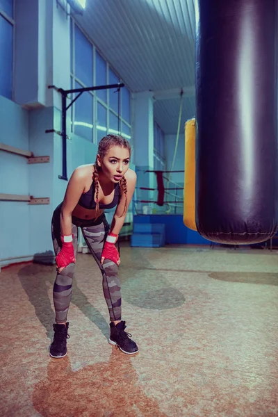 Portrait of female boxer in sport wear with fighting stance against spotlight. Sexy fitness blonde girl in sport wear with perfect body in boxing gym, posing. Sport, fitness, lifestyle concept