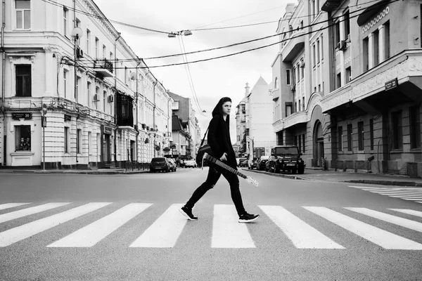 Monochrome photo. Guitarist in dark clothes crosses the street on the transition