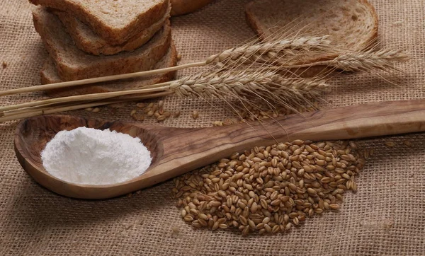 Öronen av vete, frön, bröd, rag och mjöl i trä sked på lin, jute bakgrund, textur — Stockfoto