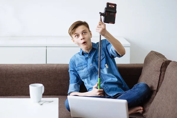 Şaşırmış genç adam arkadaşlarıyla uzaktan iletişim kuruyor ve evdeki oturma odasında kanepede otururken video konferansında eğleniyor. Selfie çubuğu.
