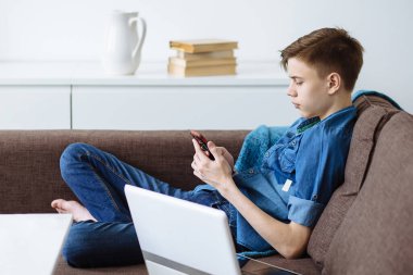 Düşünceli genç adam oturma odasındaki kanepede dizüstü bilgisayarın yanında otururken beyaz mobilya ve kitapların arka planında cep telefonu ekranına bakıyor. Çevrimiçi iletişim, online eğitim.