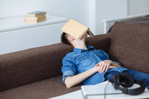 Young guy student fell asleep with book on his face on sofa in living room. Student was tired and fell asleep. Exam preparation.