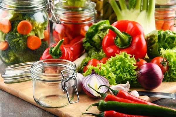 Frascos con comida marinada y verduras crudas en la tabla de cortar — Foto de Stock