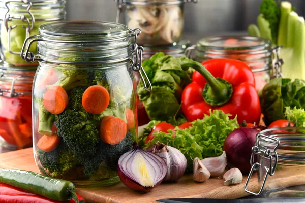 Frascos con comida marinada y verduras crudas en la tabla de cortar — Foto de Stock