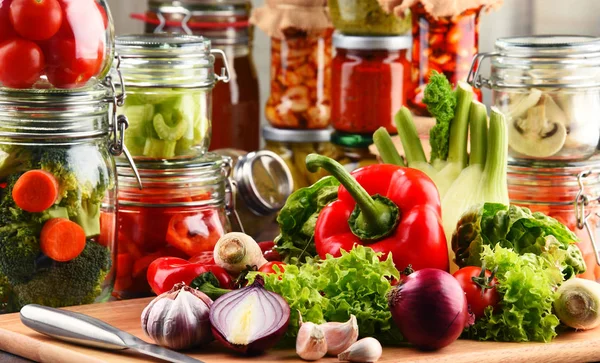 Frascos con comida marinada y verduras crudas en la tabla de cortar — Foto de Stock