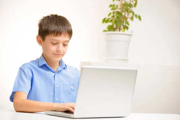 Niño en una camisa azul utiliza un ordenador portátil sentado en el interior de una mesa — Foto de Stock