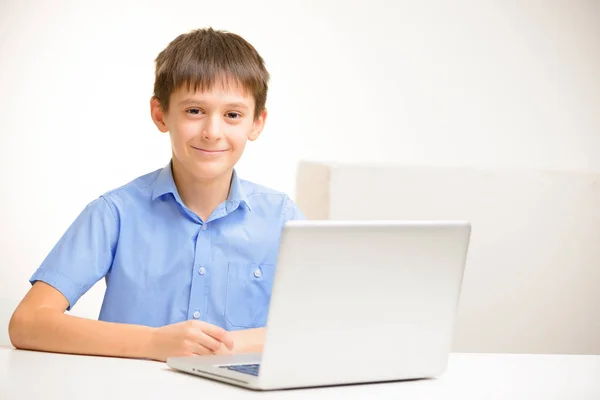 Niño en una camisa azul utiliza un ordenador portátil sentado en el interior de una mesa — Foto de Stock