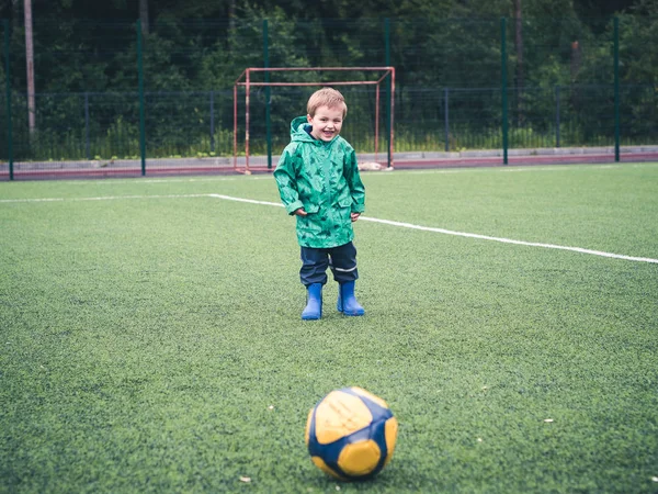 Lindo niño pequeño está jugando con una pelota de fútbol amarillo en un parque infantil. Deporte, mal tiempo, lluvia . — Foto de Stock