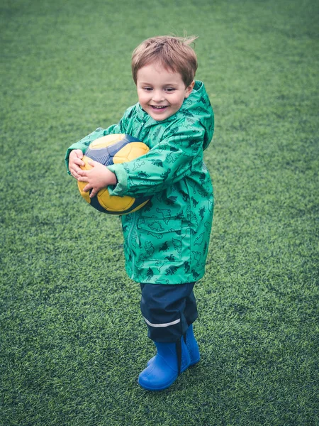 Lindo niño pequeño está jugando con una pelota de fútbol amarillo en un programa de juegos para niños. Deporte, mal tiempo, lluvia . — Foto de Stock