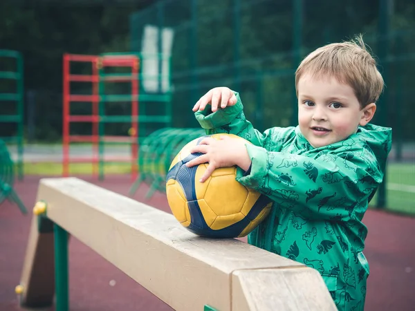 Lindo niño pequeño está jugando con una pelota de fútbol amarillo en un parque infantil. Deporte, mal tiempo, lluvia . — Foto de Stock