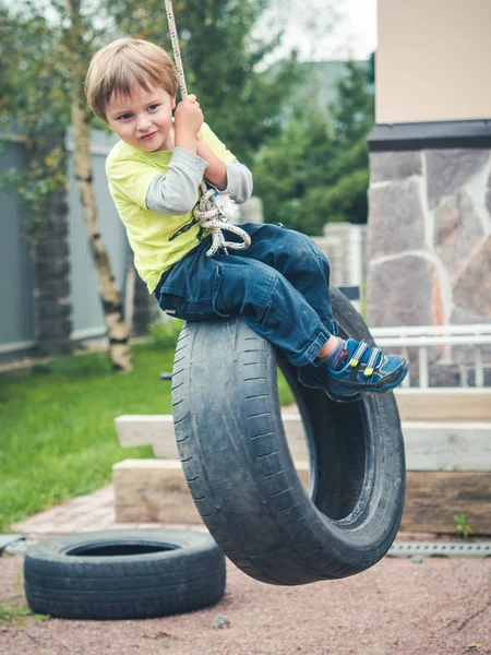 Niño feliz balanceándose sobre un neumático usado como columpio. Concepto foto de la infancia, nostalgia, memoria, pasado, vida, retro, vendimia, hogar dulce hogar . — Foto de Stock