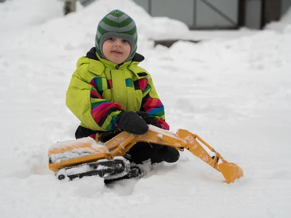 Cute cheerful child dressed in winter overalls playing excavator on white snow Royalty Free Stock Photos