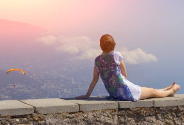 güzel bir elbise kız bir taş çıkıntıda yatıyor ve onun yanında gökyüzünde yamaç paraşütü üzerinde güzel bir gün batımı ve bir atlet bakıyor