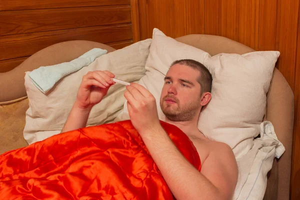 Kranker liegt mit roter Decke im Bett, in den Händen ein Thermometer — Stockfoto