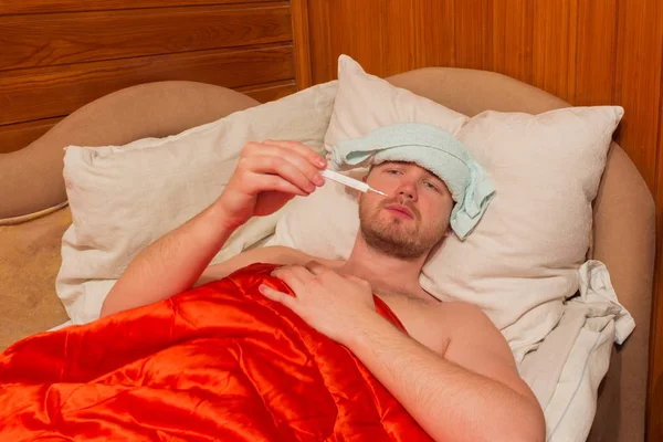 Kranker liegt mit roter Decke im Bett, in den Händen ein Thermometer — Stockfoto
