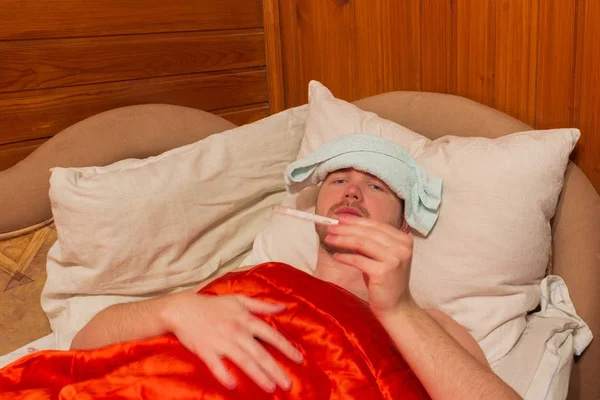 Kranker liegt mit roter Decke im Bett, in den Händen ein Thermometer — Stockfoto