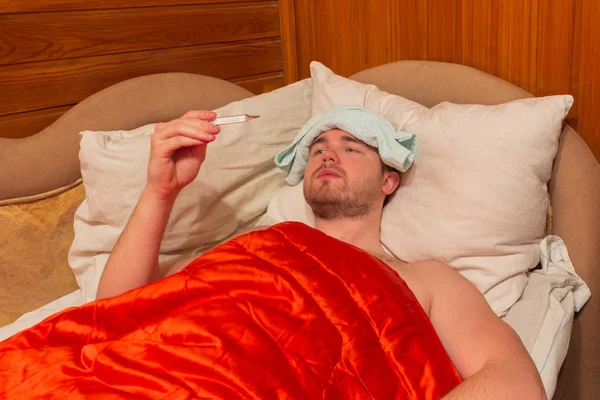 Kranker liegt mit roter Decke im Bett, in den Händen ein Thermometer — Stockfoto