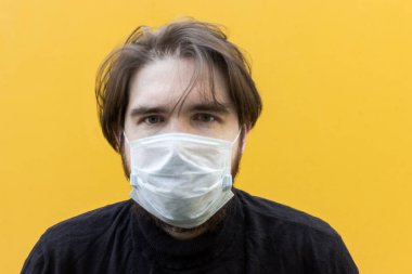 Koronavirüsten korunmak için tek kullanımlık maske takan siyah ceketli bir adam. Sarı arka plan, yüzünde duygular. dağınık erkek saçı. Hastalık durumunda güvenlik önlemleri.