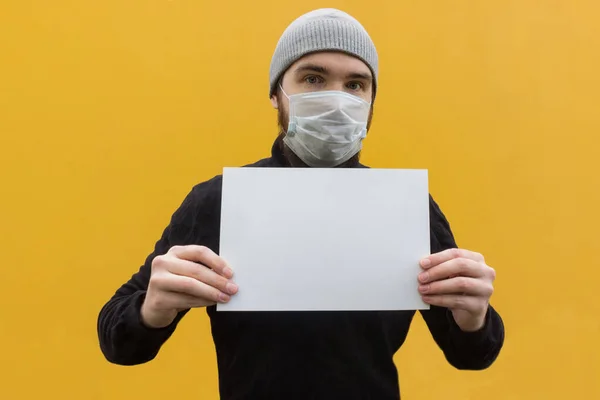 Man in a black jacket wearing a disposable mask to protect against coronavirus. Yellow background, emotions on the eyes, grey hat with empty sheet of paper in hands.Safety measures in case of illness.