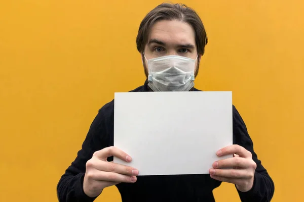 Koronavirüsten korunmak için tek kullanımlık maske takan siyah ceketli bir adam. Sarı arka plan, gözlerinde duygular, ellerinde boş kağıt tutuyor. hastalık ihtimaline karşı tedbirler..