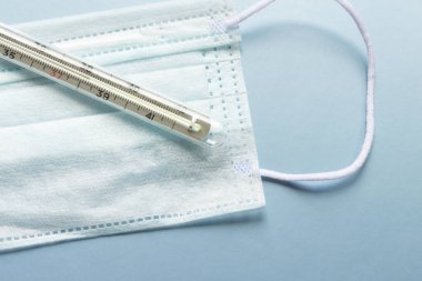 İnsanları tehlikeli bir virüsten korumak için mavi tıbbi tek kullanımlık yüz maskesi ve termometre 2020 renk eğiliminin arka planında yüksek ateş olduğunu gösteriyor. Çin 'de koronavirüs salgını için sağlık ve tıbbi konsept.