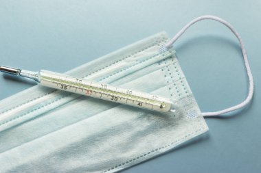 İnsanları tehlikeli bir virüsten korumak için mavi tıbbi tek kullanımlık yüz maskesi ve termometre 2020 renk eğiliminin arka planında yüksek ateş olduğunu gösteriyor. Çin 'de koronavirüs salgını için sağlık ve tıbbi konsept.