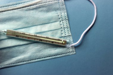 İnsanları tehlikeli bir virüsten korumak için mavi tıbbi tek kullanımlık yüz maskesi ve termometre 2020 renk eğiliminin arka planında yüksek ateş olduğunu gösteriyor. Çin 'de koronavirüs salgını için sağlık ve tıbbi konsept.