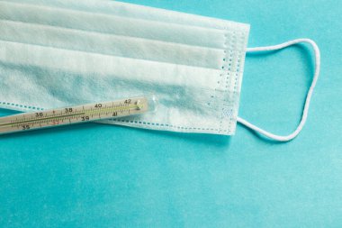 İnsanları tehlikeli bir virüsten korumak için mavi bir tıbbi tek kullanımlık maske 2020 renk eğiliminin arka planında yatıyor. Çin 'de koronavirüs salgını için sağlık ve tıbbi konsept. Termometre yüksek sıcaklık gösteriyor