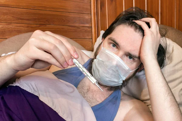 a man with dark hair and a disposable mask to protect against the virus, lies at home in bed, isolating himself from other people. fever, headache and nausea are symptoms - take care of your health