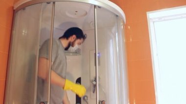 Coronavirus sırasında ev güvenliği için duş kabininin hijyenik temizliği. Gri tişörtlü ve sakallı bir adam, tek kullanımlık maske takıyor, banyoyu dezenfekte ediyor. Böyle üzücü bir zamanda güçlü bir bağışıklık sistemi.