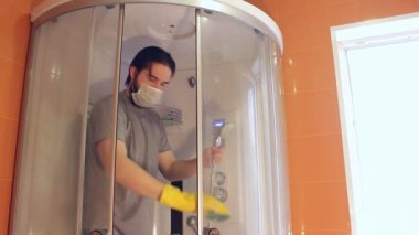 Coronavirus sırasında ev güvenliği için duş kabininin hijyenik temizliği. Gri tişörtlü ve sakallı bir adam, tek kullanımlık maske takıyor, banyoyu dezenfekte ediyor. Böyle üzücü bir zamanda güçlü bir bağışıklık sistemi.