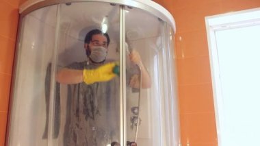Coronavirus sırasında ev güvenliği için duş kabininin hijyenik temizliği. Gri tişörtlü ve sakallı bir adam, tek kullanımlık maske takıyor, banyoyu dezenfekte ediyor. Böyle üzücü bir zamanda güçlü bir bağışıklık sistemi.