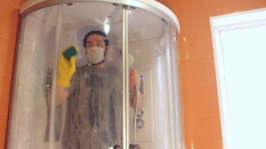Coronavirus sırasında ev güvenliği için duş kabininin hijyenik temizliği. Gri tişörtlü ve sakallı bir adam, tek kullanımlık maske takıyor, banyoyu dezenfekte ediyor. Böyle üzücü bir zamanda güçlü bir bağışıklık sistemi.