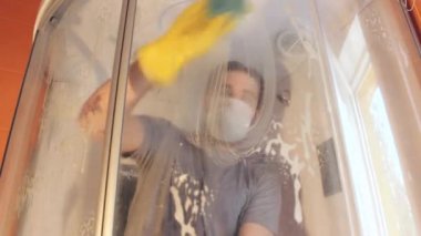 Coronavirus sırasında ev güvenliği için duş kabininin hijyenik temizliği. Gri tişörtlü ve sakallı bir adam, tek kullanımlık maske takıyor, banyoyu dezenfekte ediyor. Böyle üzücü bir zamanda güçlü bir bağışıklık sistemi.