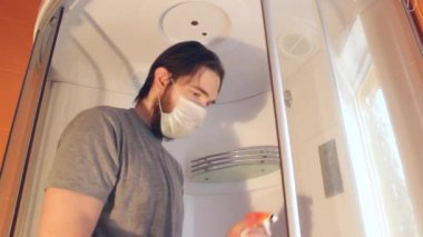Coronavirus sırasında ev güvenliği için duş kabininin hijyenik temizliği. Gri tişörtlü ve sakallı bir adam, tek kullanımlık maske takıyor, banyoyu dezenfekte ediyor. Böyle üzücü bir zamanda güçlü bir bağışıklık sistemi.