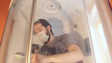 Coronavirus sırasında ev güvenliği için duş kabininin hijyenik temizliği. Gri tişörtlü ve sakallı bir adam, tek kullanımlık maske takıyor, banyoyu dezenfekte ediyor. Böyle üzücü bir zamanda güçlü bir bağışıklık sistemi.