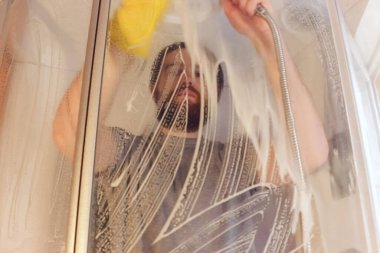 Koronavirüs sırasında güvenlik için duş kabininin hijyenik temizliği. Gri tişörtlü ve sakallı bir adam, sarı kimyasal eldiven ve temiz ajan kullanarak banyoyu dezenfekte ediyor. Bağışıklık sağlığının garantisidir.