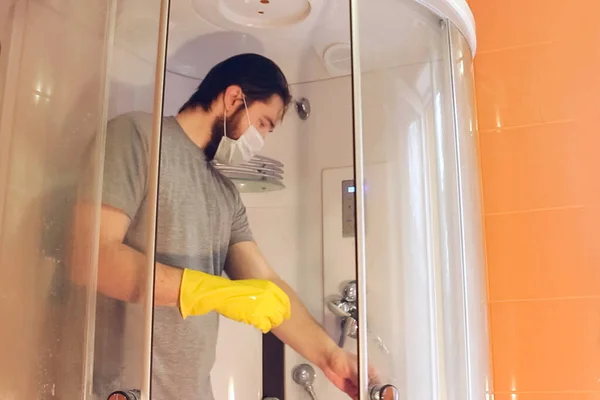 Sanitary cleaning of the shower cabin for home safety during coronavirus.A man in a gray t-shirt and beard, wearing a disposable mask, disinfects the bathroom using yellow chemical gloves and cleaning agent