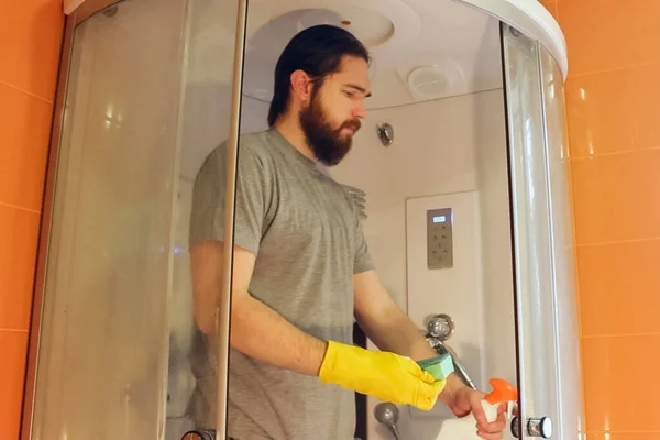 Sanitary cleaning of shower cabin for safety during coronavirus.man in a gray t-shirt and beard,disinfects the bathroom using yellow chemical gloves and clean agent.immune is the guarantee of health