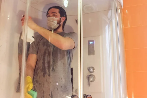 Coronavirus sırasında ev güvenliği için duş kabininin hijyenik temizliği. Gri tişörtlü ve sakallı bir adam, tek kullanımlık maske takıyor, sarı kimyasal eldiven ve temizlik malzemesi kullanarak banyoyu dezenfekte ediyor.