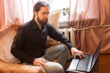 Koronavirüs sorunu yüzünden dünyada kendi kendini izole etme modu. Koyu saçlı ve kalın sakallı bir adam, evde serbest çalışan uzaktan kumandalı bir işçi olarak çalışıyor. Laptop ve telefon, kötü öfke ve korku duyguları.