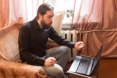 Koronavirüs sorunu yüzünden dünyada kendi kendini izole etme modu. Koyu saçlı ve kalın sakallı bir adam, evde serbest çalışan uzaktan kumandalı bir işçi olarak çalışıyor. Laptop ve telefon, kötü öfke ve korku duyguları.