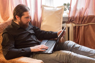 Koronavirüs sorunu yüzünden dünyada kendi kendini izole etme modu. Koyu saçlı ve kalın sakallı bir adam, evde serbest çalışan uzaktan kumandalı bir işçi olarak çalışıyor. Laptop ve telefon, kötü öfke ve korku duyguları.