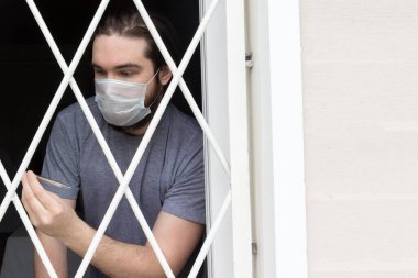 Tek kullanımlık maskeli, siyah saçlı, sakallı ve gri tişörtlü bir erkek manken. Coronavirus hastası evde karantina ve hastalık önleme altında kalıyor. Bir insanın gözlerinde olumsuz ve olumsuz duygular